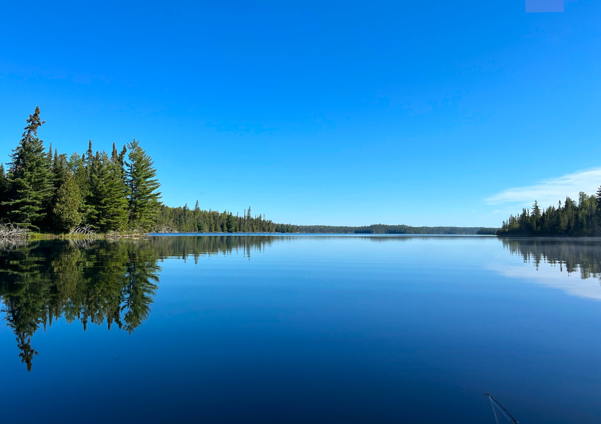 Pic of lake in Quetico with tufts of pine trees on the right and left and the left tuft trailing off in the distance. The blue sky is reflected on the lake, with a smattering of white clouds just above the trees on the right. Peaceful and quiet.